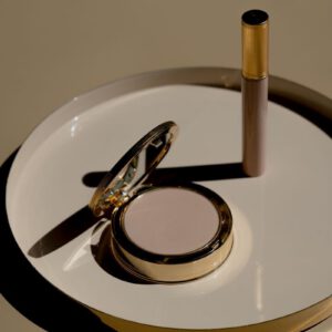 Golden makeup compact with matching mascara on beige tray, representing luxury cosmetics.